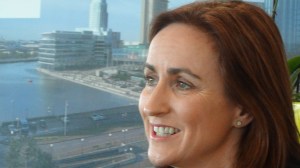 Carmel Booth of Atlantic Gateway, with a view of Liverpool's Mersey waterfront in the background: a road with lampposts and trees, parked vehicles, major buildings and a small boat on the wide river.